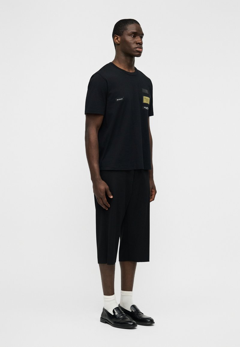 T-shirt en coton noir à coupe décontractée, associé à un short noir. Détails remarquables : un patch sur la poitrine et des mocassins noirs portés avec des chaussettes blanches.