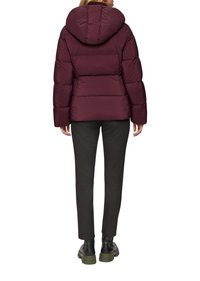 Doudoune bordeaux avec capuche, design matelassé et poignets élastiques. Associée à un pantalon noir ajusté et des bottes noires imposantes.