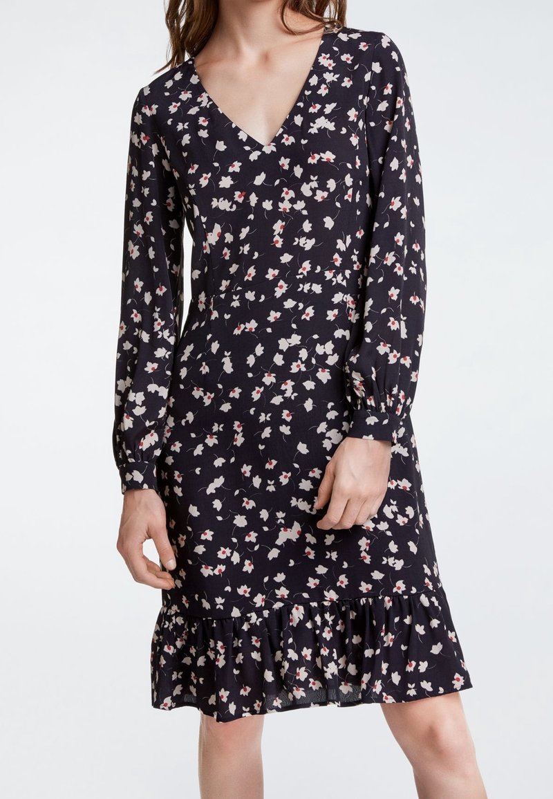 Robe fleurie en marine avec des manches longues, décolleté en V et ourlet volanté. Présente de petits motifs floraux blancs et rouges sur un fond sombre.