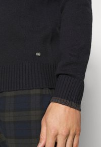 Mörkblå stickad tröja med ribbad nederkant och ärmslut, med en liten silverlogotyp ovanför nederkanten. Bärs över byxor med rutigt mönster.