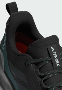 Schwarzer Sportschuh mit strukturiertem Obermaterial, verstärktem Fersenbereich und grünen Akzenten. Verfügt über einen gepolsterten Kragen und reflektierende Schnürsenkel. Logo an der Seite angebracht.