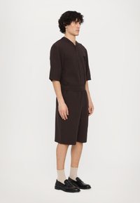 Dark brown short-sleeve shirt with a button front and a chest pocket, paired with matching knee-length shorts. Black loafers and beige socks.