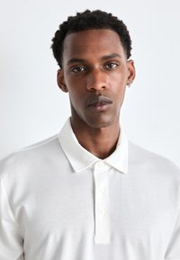 Bílá polo košile s klasickým límcem, obsahující klopu se třemi knoflíky a texturovanou látku. Model má krátké, kudrnaté vlasy.