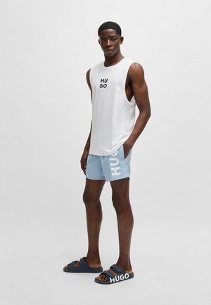 White sleeveless tank top with black "HUGO" logo, light blue shorts featuring a white "HUGO" print, dark sandals with "HUGO" branding.