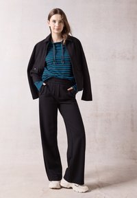 Schwarze Jacke über einem teal-gestreiften Hoodie, kombiniert mit schwarzen weit geschnittenen Hosen und weißen Sneakers. Glatte Stoffoberflächen und lässige Passform.