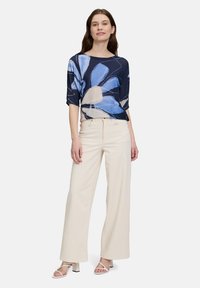Granatowa bluzka w kwiatowy wzór z krótkimi rękawami, zestawiona z beżowymi spodniami w szerokiej nogawce i białymi sandałami na obcasie. Proste tło.