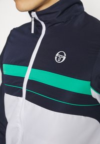 Navy zip-up jacket featuring a white and green horizontal stripe across the chest, textured fabric, and a circular logo on the left side.