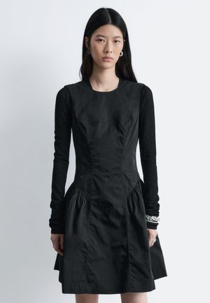 Sort kjole med tætsiddende overdel, voluminøst skørt og lange teksturerede ærmer. Har rund halsudskæring og strukturerede syninger.