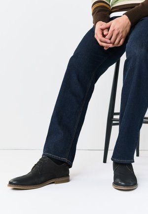Personne assise portant un pull rayé, un jean bleu foncé et des chaussures en cuir noires à lacets, assise sur un tabouret noir sur fond blanc.