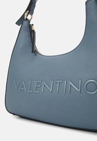 Φωτεινό μπλε δερμάτινη τσάντα χεριού με καμπύλη μορφή, που διαθέτει ανάγλυφο λογότυπο "VALENTINO" και χρυσές λεπτομέρειες σε φερμουάρ. Λείο υλικό με αντιθετικά άκρα.