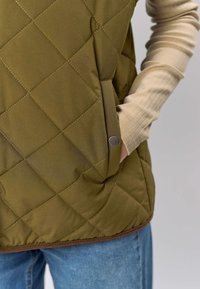 Quiltet vest i olivengrønn med en teksturert finish, sidelommer og metallknappedetaljer, kombinert med en ribbestrikket beige langermet skjorte.