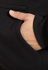 Materiale di felpa nera con una tasca frontale. In primo piano si vede una mano appoggiata nella tasca, con polsini a costine e dettagli di cucitura ordinati.
