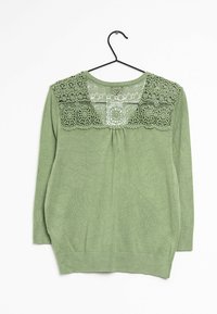 Vaaleanvihreä pitkähihainen neule, jossa on kukkakuvioinen pitsipaneeli yläselässä, roikkumassa mustassa langassa valkoista taustaa vasten.