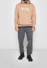 Felpa beige con scritta "NEW YORK", abbinata a jeans grigi con effetto usurato e sneaker nere. Vestibilità casual con dettaglio della tasca frontale.