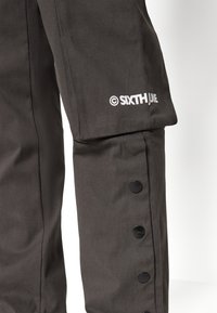 Mørkegrå bukser med et elegant design, som har svarte trykknappdetaljer langs siden og en hvit logo på kanten.