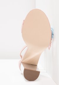 Vue de la semelle d'une sandale rose à talon haut avec un talon carré et des décorations florales sur une surface blanche contre un mur blanc.