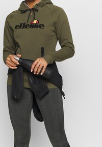 Olijfgroene hoodie met zwart 'ellesse' logo, koordjes voor de capuchon en ritszakken. Gecombineerd met donkergrijze legging en een zwarte waterfles.