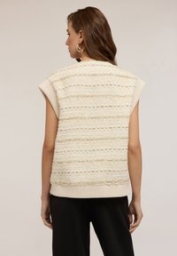Donna con capelli castani mossi indossa un gilet in maglia beige senza maniche e pantaloni neri, in piedi di spalle contro uno sfondo semplice.