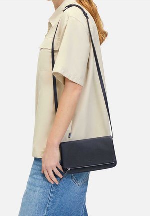 Femme portant une chemise beige à manches courtes et un jean bleu, tenant un sac bandoulière noir avec une fermeture éclair.