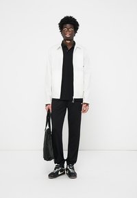 Witte lichtgewicht jas over een zwart poloshirt, gecombineerd met zwarte broek en zwarte sneakers. Houdt een zwarte tas vast. Minimalistisch ontwerp en kleurenschema.