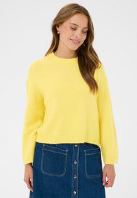 Pull jaune en maille avec un col rond et des manches longues ; texture douce. Porté avec une jupe en denim ornée de boutons et de poches.