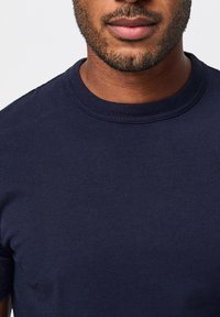 Kragenloses navyblaues Baumwoll-T-Shirt mit kurzen Ärmeln; glatte Textur und minimal sichtbare Nähte am Halsausschnitt.