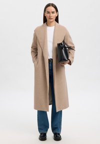 Kvinne som har på seg en lang beige frakk, hvit skjorte, blå jeans, svarte sko, holder en svart håndveske, står mot en ensfarget lys bakgrunn.