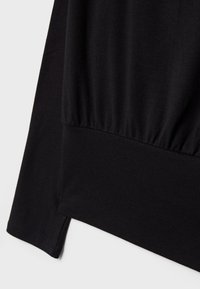 Manche et ourlet inférieur d'une chemise noire à manches longues avec un détail de tissu froncé sur un fond clair uni.