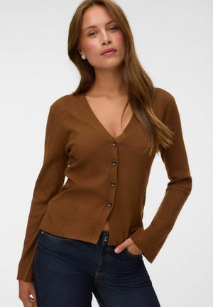 Femme aux longs cheveux châtain clair, portant des boucles d’oreilles en perles, un cardigan marron boutonné et un jean bleu foncé, posant avec une main dans la poche.