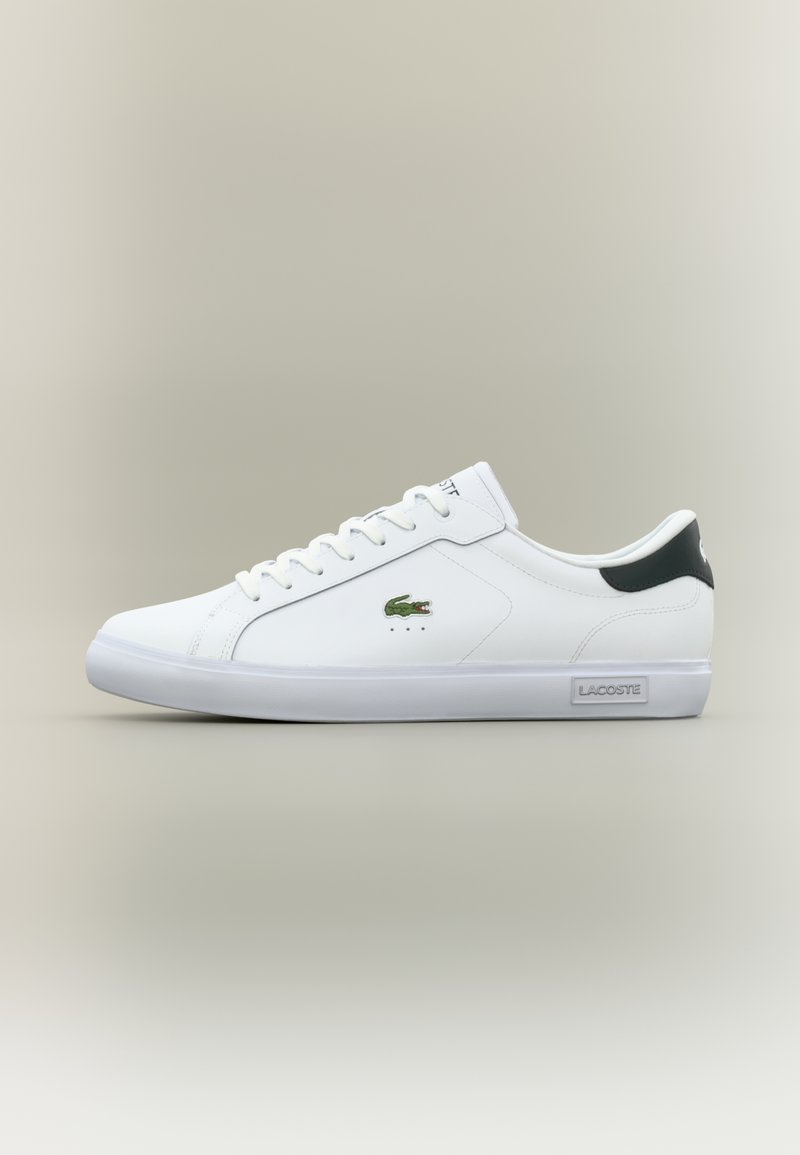 Vita låga sneakers med grönt krokodil-logotyp på sidan, svart hälflik, vita snören och "Lacoste"-etikett på sula.