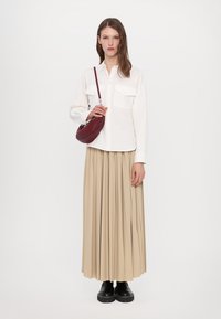 Chemise blanche à boutons avec deux poches avant, jupe plissée beige et bottes noires à semelles épaisses ; portant un petit sac à bandoulière bordeaux.