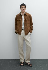 Brun mocka skjortjacka med två bröstfickor, vit skjorta under, beige byxor och svarta sandaler med remmar. Enfärgad bakgrund.