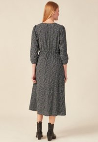 Schwarzes Blumenkleid in Midi-Länge mit lila und grünen Mustern, elastischem Bund, Dreiviertelärmeln und einem flatternden Rock. Getragen mit schwarzen ankle boots.