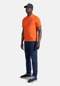 Oranžové polo tričko s logem, námořnické kalhoty a námořnická čepice. Tričko má límeček a krátké rukávy; čepice má kšilt a detaily loga.