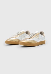 Sneakers med en skinnende sølv- og gull læroverdel, beige detaljer og en teksturert gummisåle. Har lisser og merkevare på siden.