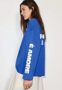 Blauw katoenen shirt met lange mouwen, met opvallende witte tekst op de mouwen. Het ontwerp heeft een losse pasvorm, een ronde halslijn en een effen achtergrond.