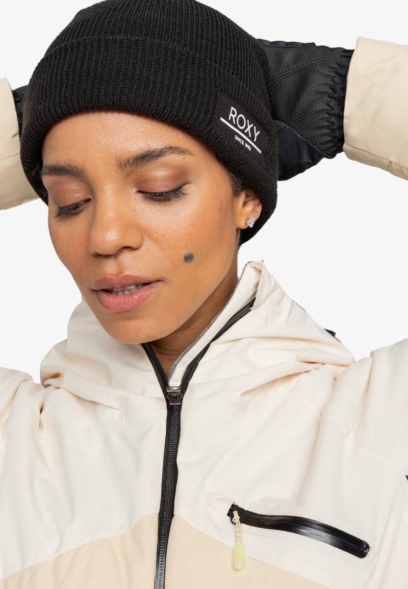 Svart strikket lue med logo; myk tekstur. Iført en kremfarget og beige zip-up jakke med hette og sidelommer. Fokus på ansikt og tilbehør.