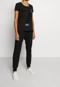 Musta t-paita pyöreällä kaula-aukolla, jossa on teksturoitu logo ja graafinen suunnittelu. Yhdistetty mustiin verkkareihin ja valkoisiin lenkkikenkiin.