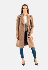 Abrigo largo en tono beige, hecho de tela texturizada con cuello con muesca, cierre de un solo botón y bolsillos laterales. Llevado sobre una camisa clara y jeans.