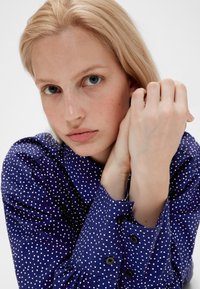 Blue polka dot blouse with a button-down collar, gathered sleeves, and black button accents, featuring a soft textured fabric.