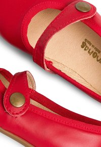 Zapatos de cuero rojo con puntera redondeada, correa elástica y cierre metálico a presión. El interior presenta una textura de cuero en tono beige claro.