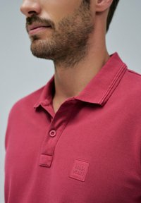 Primer plano de un hombre que lleva una camisa polo de textura en color rosa oscuro con un parche del logo "Salsa Jeans" en el pecho.