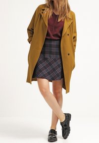 Senapsgul överdimensionerad kappa, vinröd topp, rutiga kjol i marinblå och gröna nyanser, svarta lackskor med en lätt glansig finish.