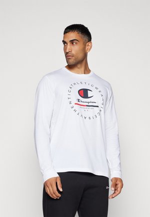 Camiseta blanca de manga larga con un logo gráfico circular en azul y rojo en el centro. Tejido suave, cuello redondo y corte relajado. Combinada con pantalones negros.