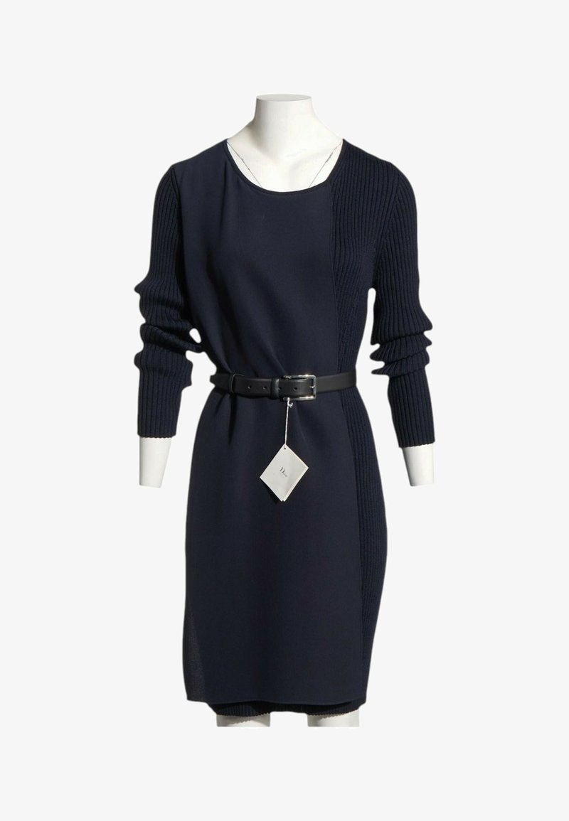 Robe en maille bleu marine avec manches côtelées et panneau latéral, ceinture noire à la taille, présentée sur un mannequin avec une étiquette blanche suspendue à la ceinture.