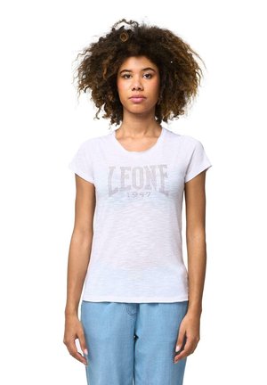 Une femme aux cheveux bouclés et volumineux se tient devant un fond blanc. Elle porte un T-shirt Leone blanc et un jean bleu clair.
