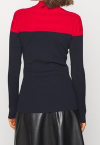 Pull en tricot présentant une moitié supérieure à rayures horizontales rouges et une moitié inférieure bleu marine foncé, avec une coupe ajustée et une texture côtelée.