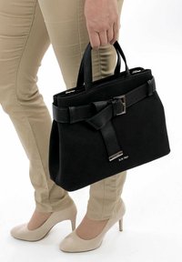 Personne portant un pantalon beige et des talons crème, tenant un sac à main noir avec un détail de boucle et des poignées courtes.