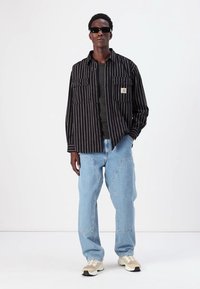 Black and white striped shirt, two chest pockets, worn over a dark shirt, paired with light blue baggy jeans and beige sneakers. Black sunglasses.