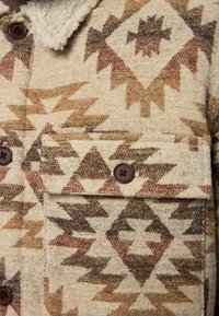 Veste en laine avec un motif géométrique crème, marron et beige. Dotée d'un col en fourrure et de deux poches poitrine avec des boutons marron.